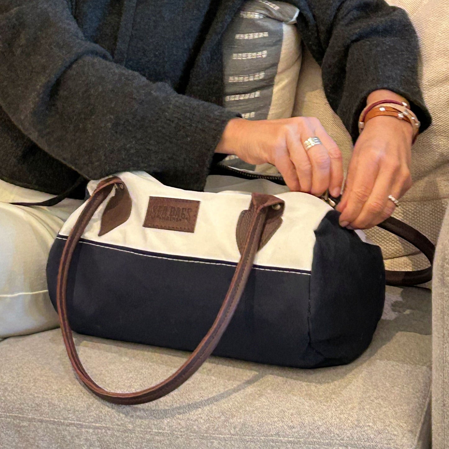 A person sits on a couch, unzipping the Sea Bags Chebeague Baguette Bag, which is made from blue and white recycled sailcloth and features brown handles. by Sea Bags