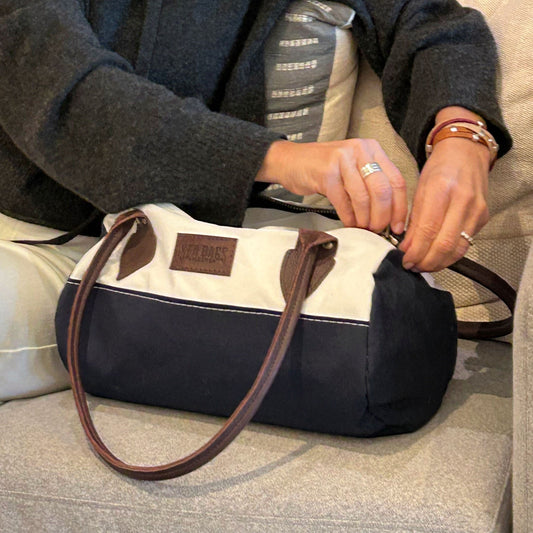 A person sits on a couch, unzipping the Sea Bags Chebeague Baguette Bag, which is made from blue and white recycled sailcloth and features brown handles. by Sea Bags