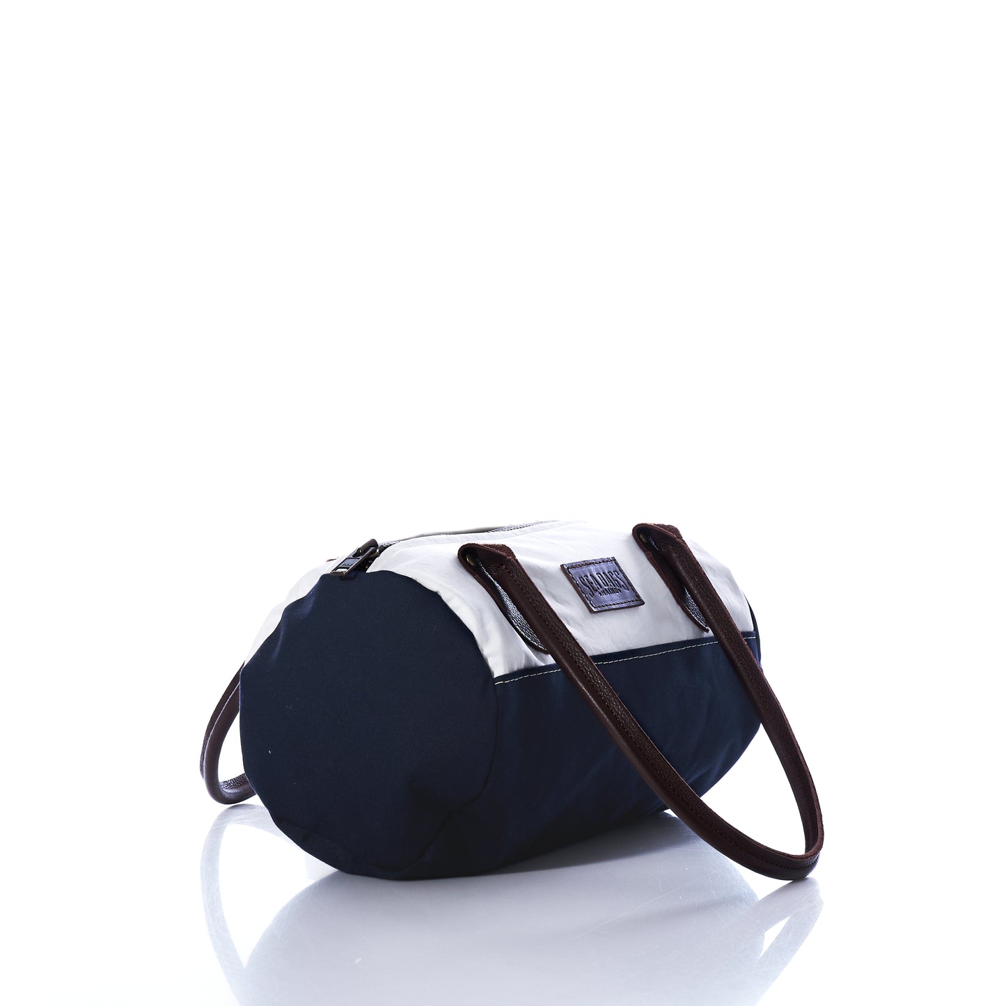 The Sea Bags Chebeague Baguette Bag, featuring a small cylindrical silhouette with black and white panels, brown handles, and a zipper top, is displayed on a white reflective surface. by Sea Bags