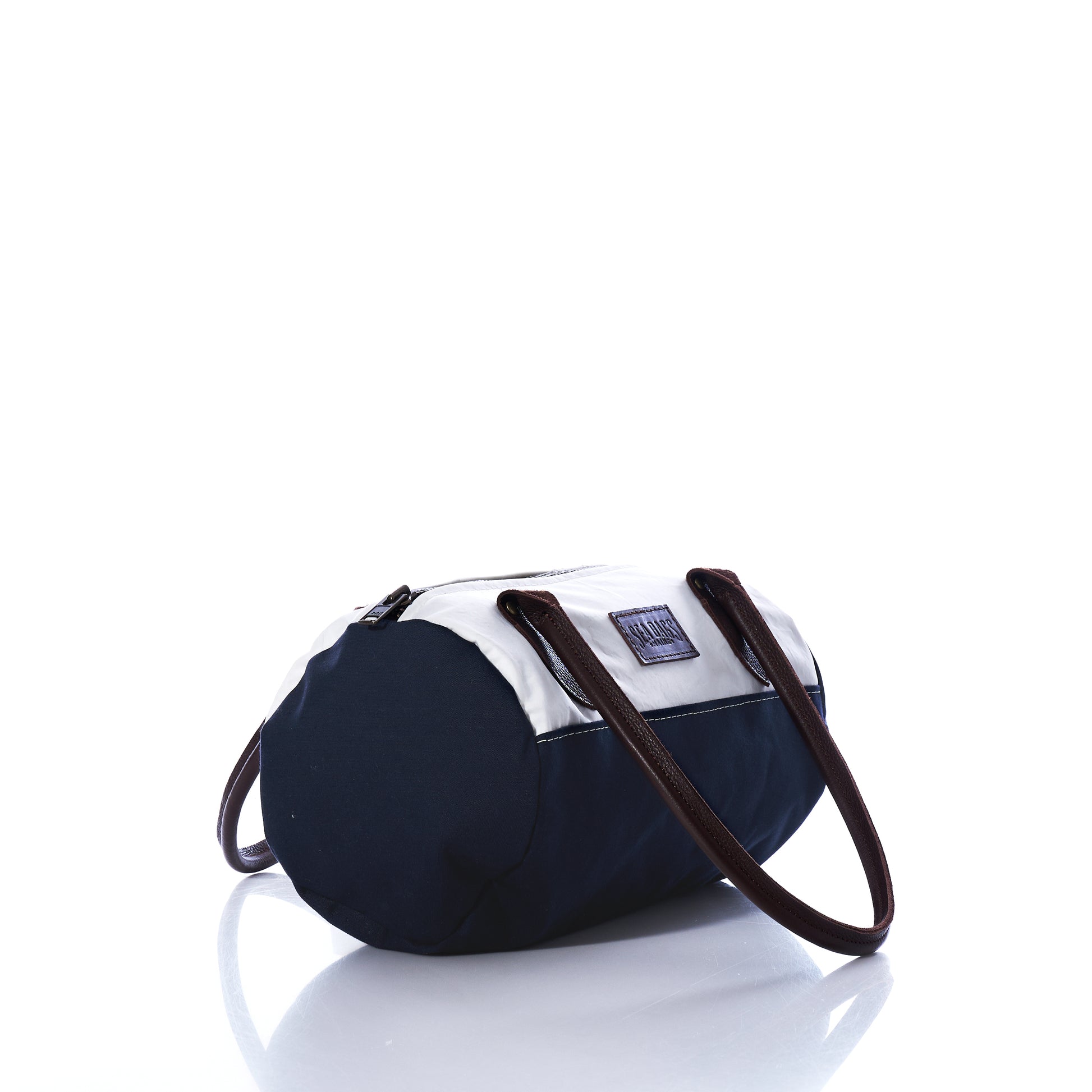 The Sea Bags Chebeague Baguette Bag, featuring a small cylindrical silhouette with black and white panels, brown handles, and a zipper top, is displayed on a white reflective surface. by Sea Bags
