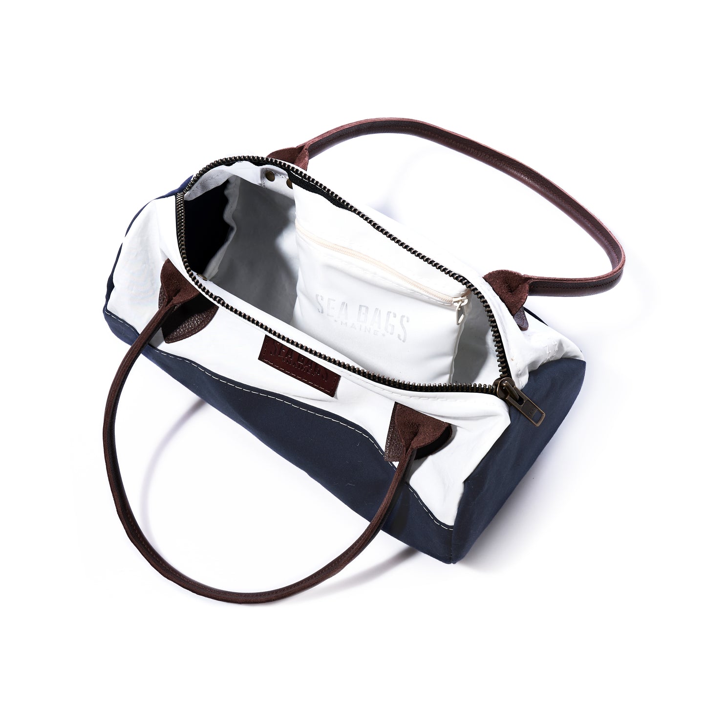 Sea Bags Chebeague Baguette Bag features a rectangular silhouette crafted from recycled sailcloth, with brown handles, a navy and white exterior, a zippered main opening, and an inner pocket labeled SEA BAGS visible inside. by Sea Bags