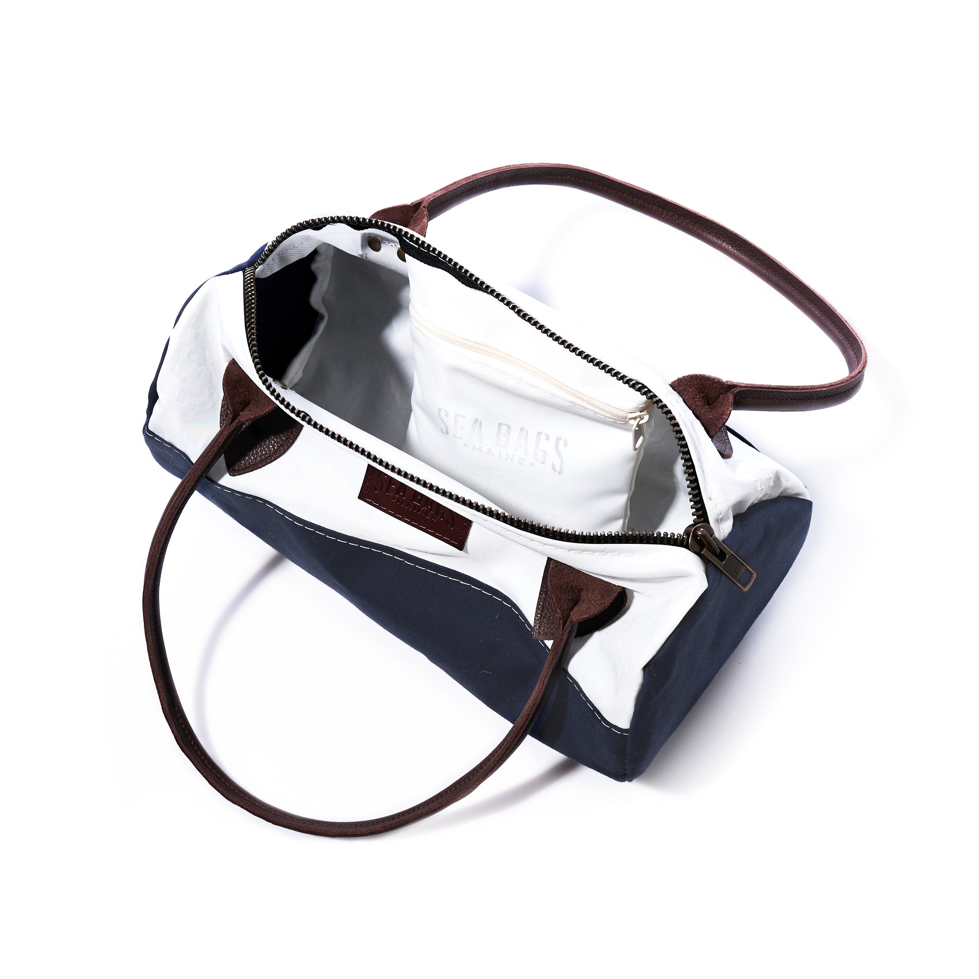 Sea Bags Chebeague Baguette Bag features a rectangular silhouette crafted from recycled sailcloth, with brown handles, a navy and white exterior, a zippered main opening, and an inner pocket labeled SEA BAGS visible inside. by Sea Bags