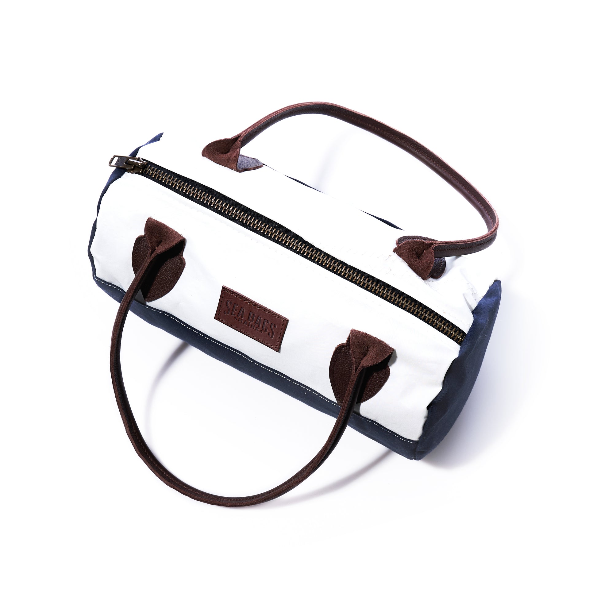 The Chebeague Baguette Bag by Sea Bags is a small cylindrical white and navy duffel bag made from recycled sailcloth, featuring brown handles and zipper, a rectangular SEA BAGS logo patch on the side, and is photographed on a white background. by Sea Bags