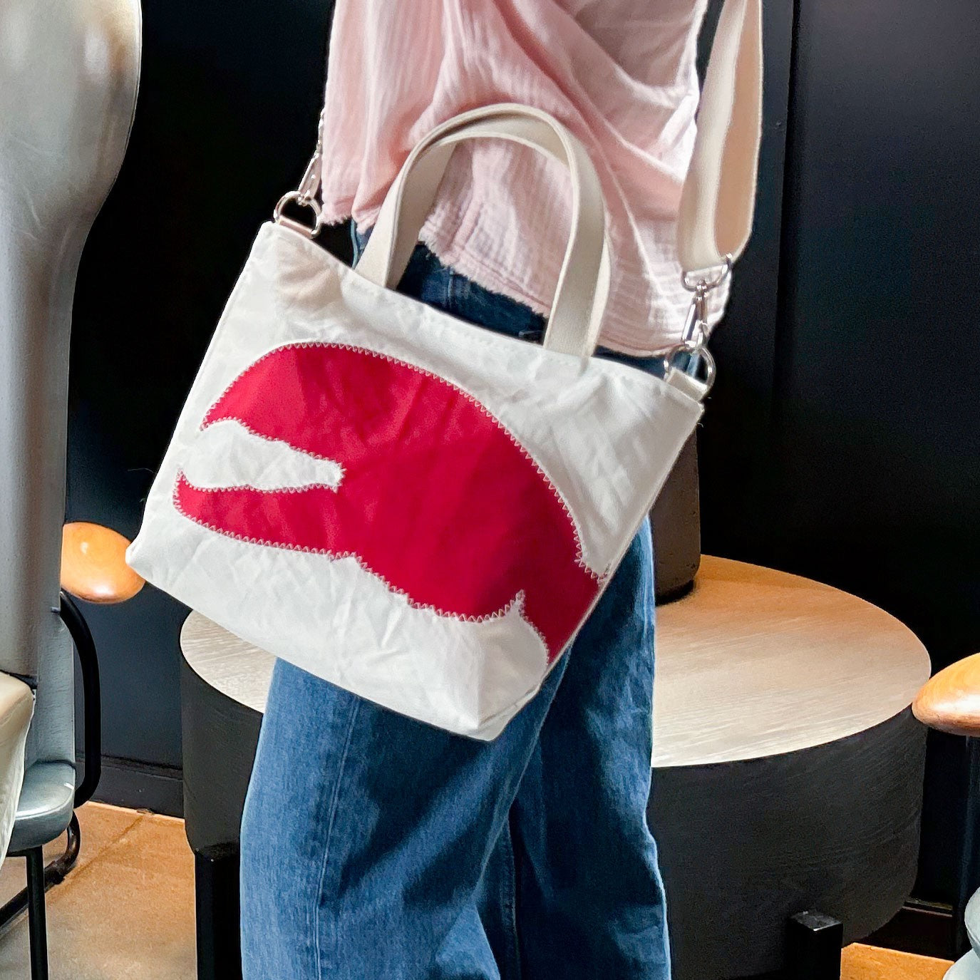 Person wearing blue jeans and a light pink top carries the Sea Bags Red Lobster Claw Crossbody Tote—an eco-friendly white bag made from recycled sailcloth, featuring a large red lobster claw design, with both short handles and a shoulder strap. by Sea Bags