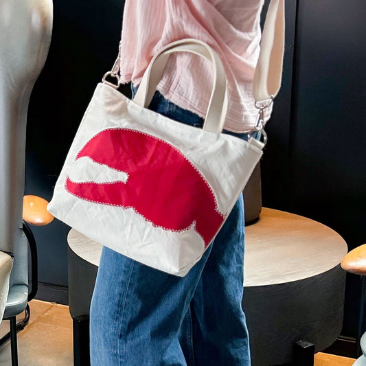 Person wearing blue jeans and a light pink top carries the Sea Bags Red Lobster Claw Crossbody Tote—an eco-friendly white bag made from recycled sailcloth, featuring a large red lobster claw design, with both short handles and a shoulder strap. by Sea Bags