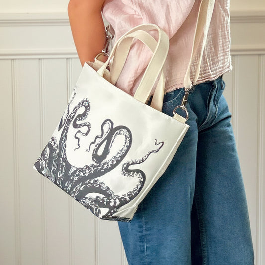 Wearing blue jeans and a light pink shirt, a person carries the Sea Bags Octopus Crossbody Tote—an eco-friendly white handbag adorned with a black octopus tentacle design and crafted from recycled sailcloth. by Sea Bags