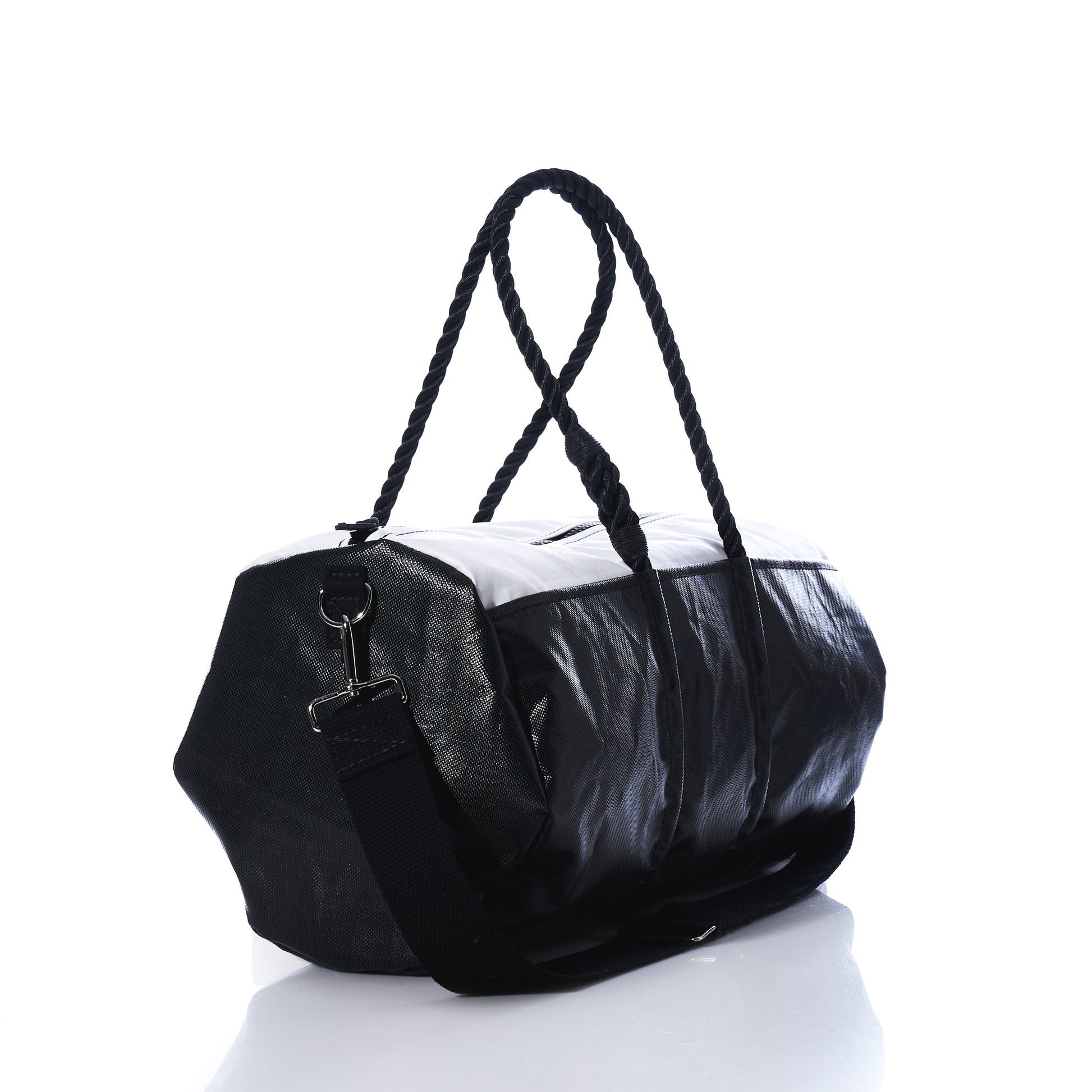 The Sea Bags Black Dragon Duffel, crafted from water-resistant recycled sail cloth, features rope handles and an adjustable shoulder strap. Photographed on a white background with a light reflection, this black and white travel bag combines durability with distinctive maritime style. by Sea Bags
