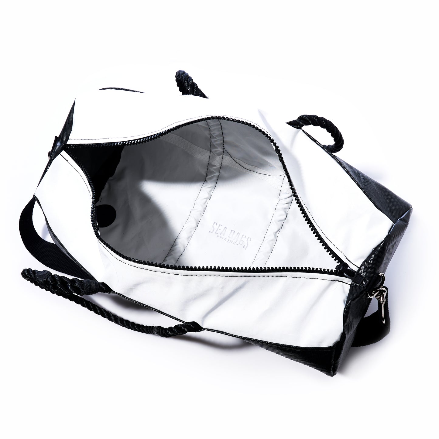 The Black Dragon Duffel by Sea Bags is a stylish travel duffel bag in black and white, featuring a rope handle. Shown unzipped and empty, it reveals a white interior crafted from recycled sail cloth with the SEA BAGS logo inside. This water-resistant bag is perfect for all your adventures. by Sea Bags