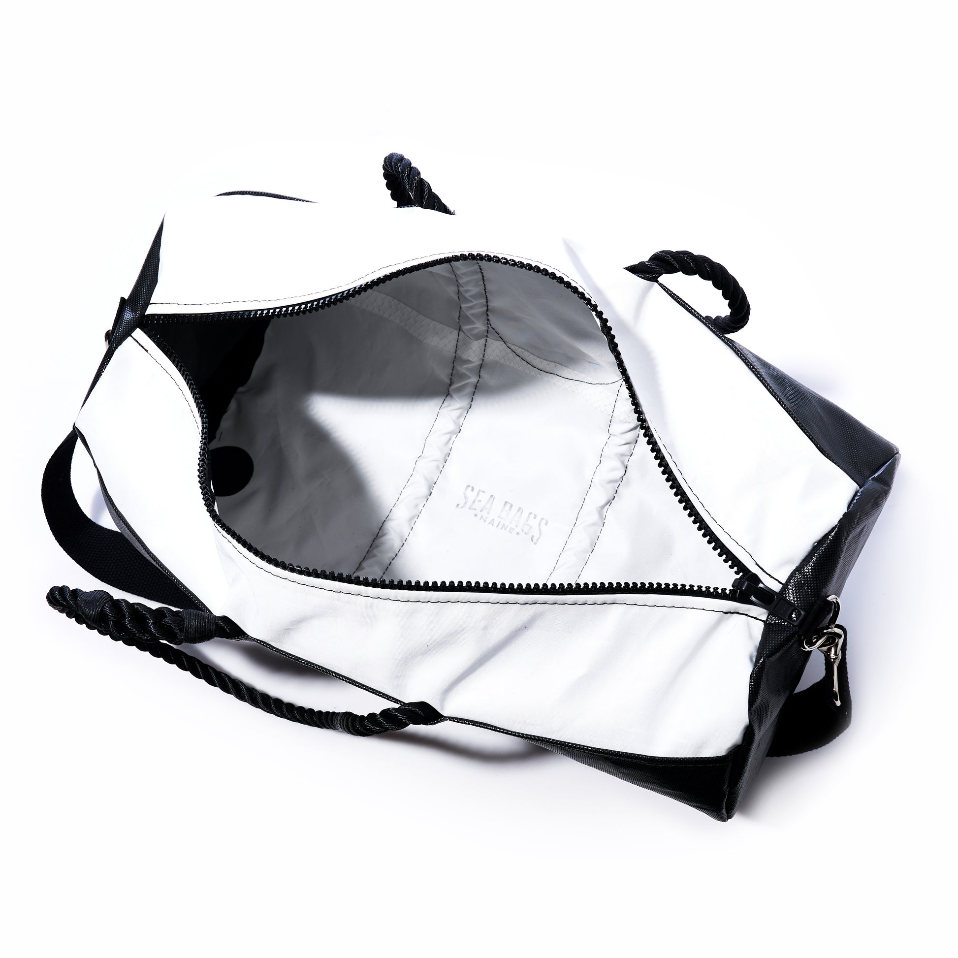 The Black Dragon Duffel by Sea Bags is a stylish travel duffel bag in black and white, featuring a rope handle. Shown unzipped and empty, it reveals a white interior crafted from recycled sail cloth with the SEA BAGS logo inside. This water-resistant bag is perfect for all your adventures. by Sea Bags