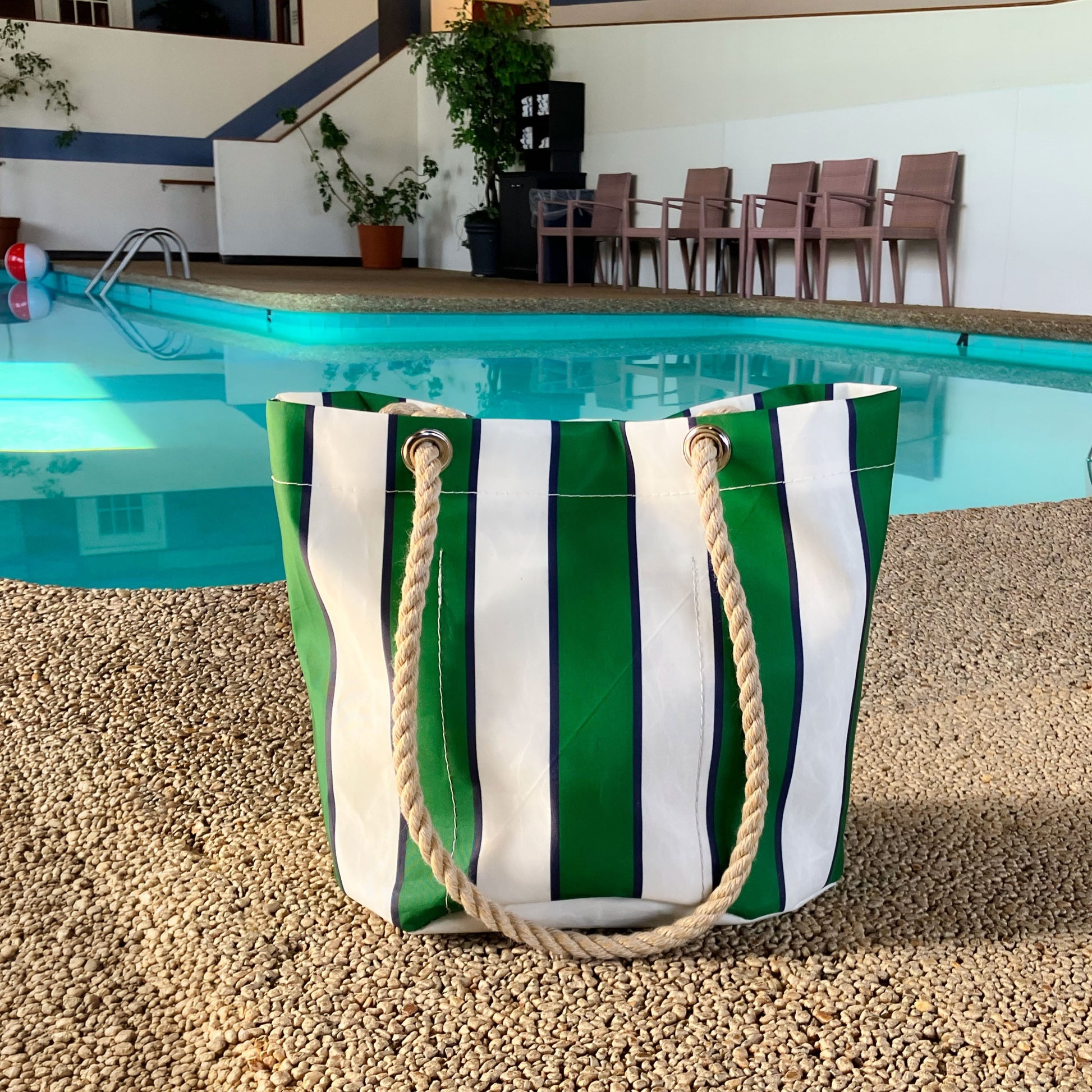 The Sea Bags Preppy Stripe Essential Handbag, featuring classic collegiate green and white stripes and sturdy rope handles, crafted from recycled sailcloth, rests on gravel near an indoor swimming pool with chairs in the background. by Sea Bags