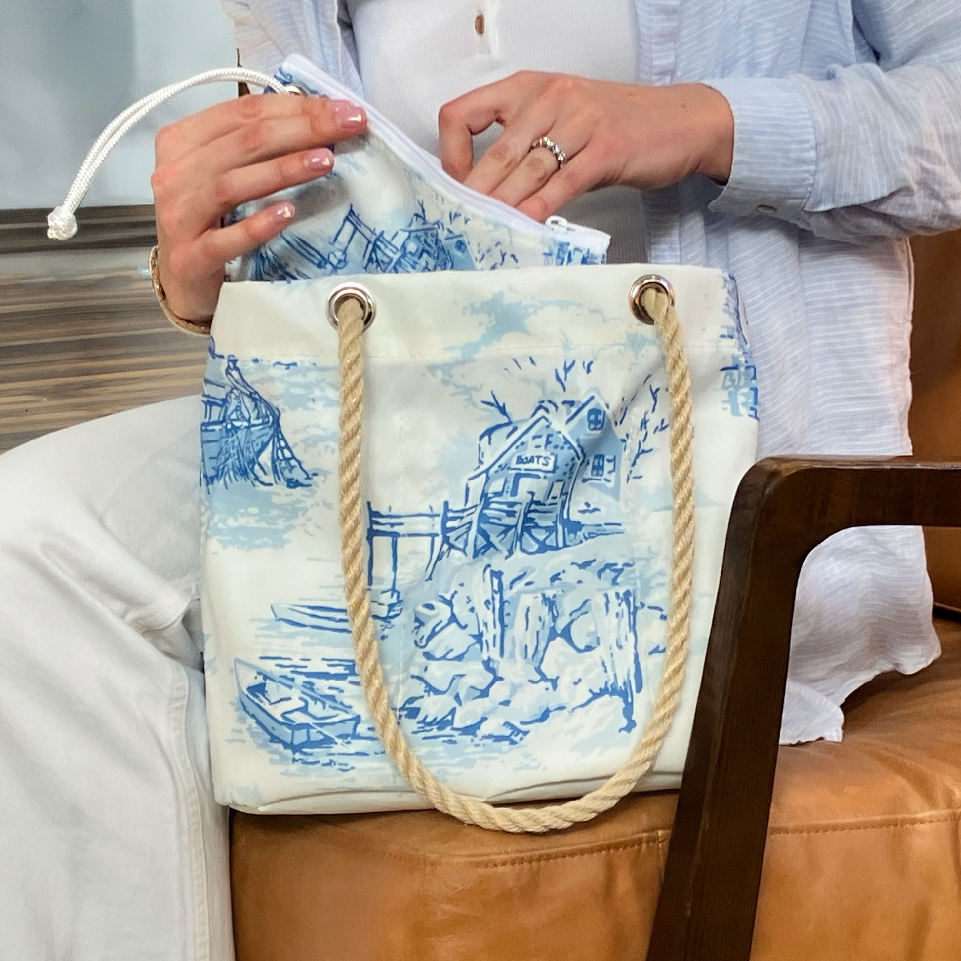 A person places the white zippered pouch into the Sea Bags Maine Fishing Village Essential Handbag—a white tote adorned with blue nautical designs and sturdy rope handles, crafted from recycled sail cloth—evoking the breezy charm of the Maine coast while sitting on a brown chair. by Sea Bags