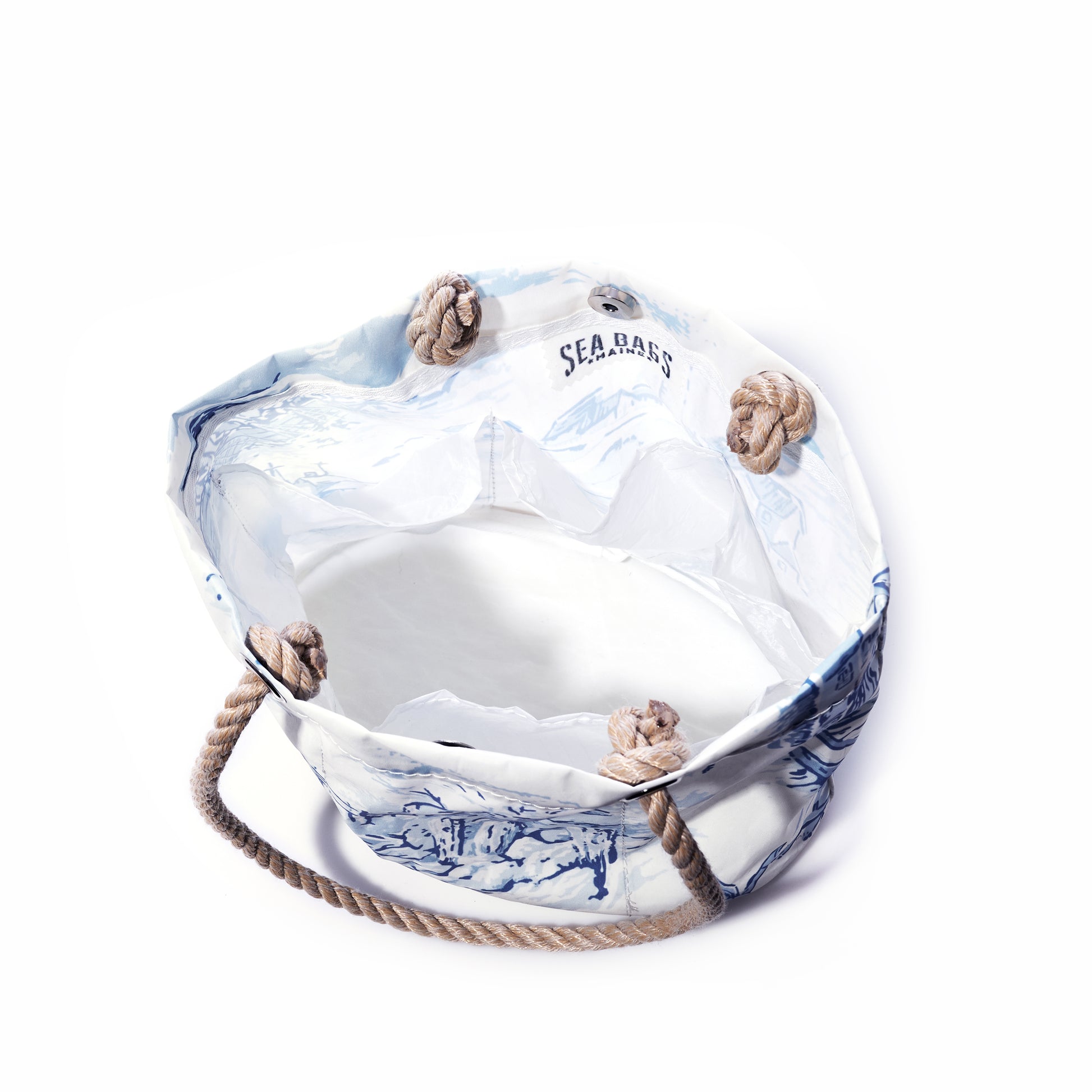 The Maine Fishing Village Essential Handbag by Sea Bags, crafted from recycled sail cloth and featuring white and blue canvas with rope handles and knotted corners, is inspired by the Maine coast. The SEA BAGS label is displayed on the inner lining, and the bag is photographed against a white background. by Sea Bags