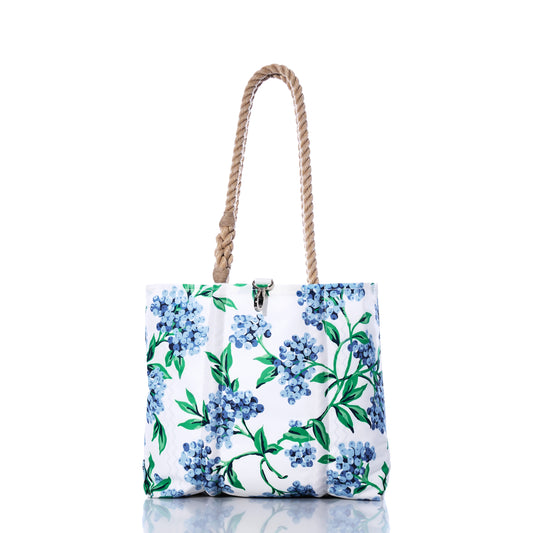 The Blueberry Print Handbag by Sea Bags showcases a blue and green floral motif inspired by Maine blueberries, is crafted from recycled sail cloth, and features two braided rope handles—its versatile design is beautifully displayed on a reflective white surface. by Sea Bags