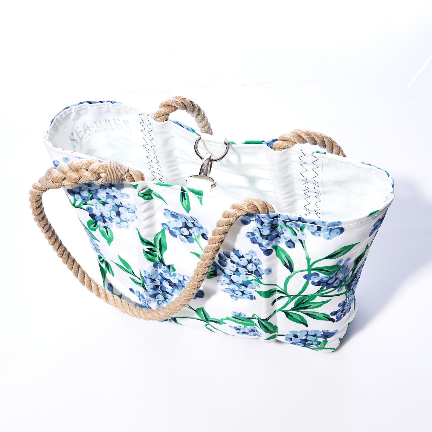 Introducing the Blueberry Print Handbag by Sea Bags—a white canvas tote adorned with a blue floral pattern inspired by Maine blueberries. This eco-friendly and versatile handbag is crafted from recycled sail cloth, features tan rope handles and a metal clasp closure, and is showcased against a white background—perfect for everyday use. by Sea Bags