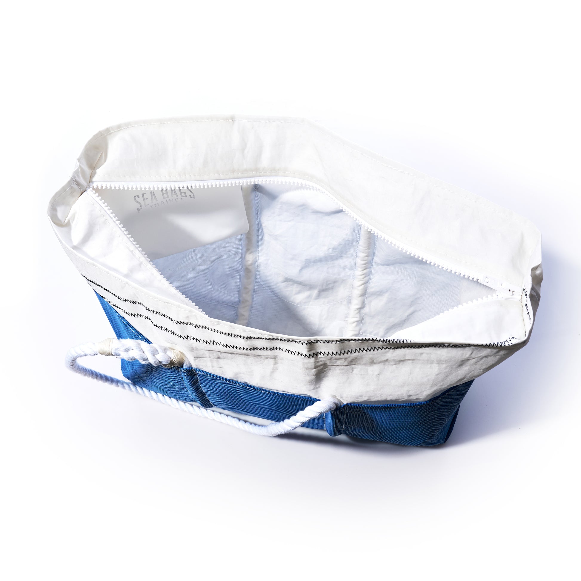 The Sea Bags Harbor Blue Beachcomber Tote, made from recycled sails, features blue and white accents, rope handles, and a zippered top. It is pictured open and empty against a white background. by Sea Bags
