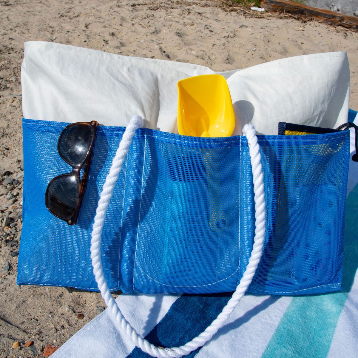 The Sea Bags Harbor Blue Beachcomber Tote, featuring white rope handles and sand-proof mesh construction, holds a white towel, a yellow plastic shovel, bottles, and sunglasses. It’s pictured resting on a beach towel at the shoreline. by Sea Bags