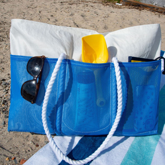 The Sea Bags Harbor Blue Beachcomber Tote, featuring white rope handles and sand-proof mesh construction, holds a white towel, a yellow plastic shovel, bottles, and sunglasses. It’s pictured resting on a beach towel at the shoreline. by Sea Bags
