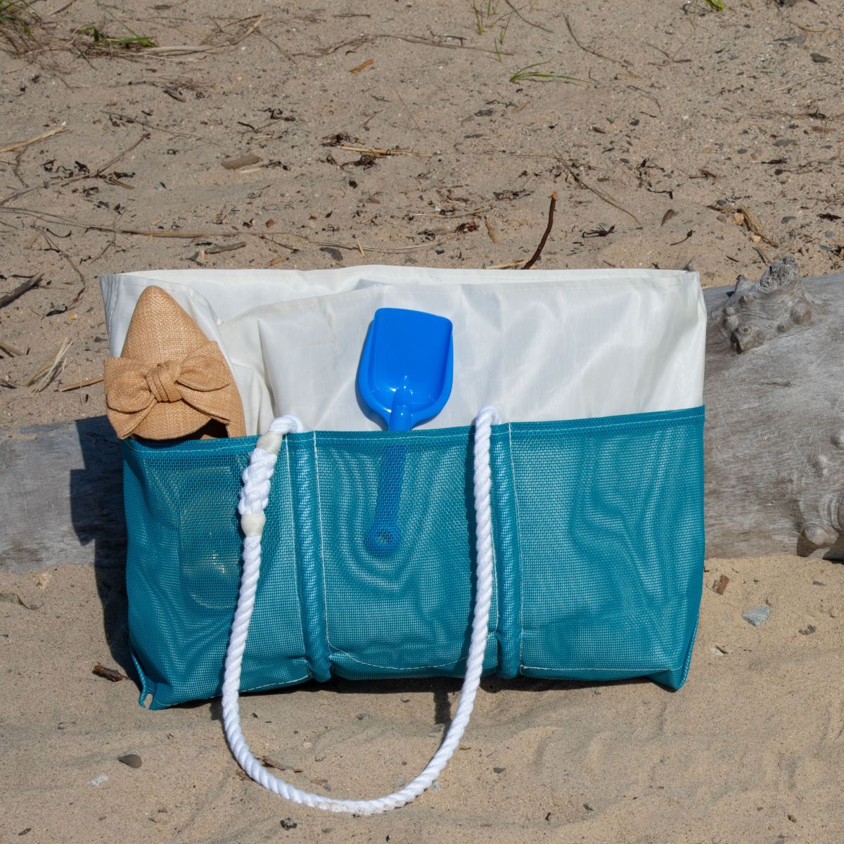 An Ocean Teal Beachcomber Tote by Sea Bags with white rope handles sits on the sand, holding a tan sunhat, a blue plastic shovel, and a white towel. A log and dry grass are in the background. by Sea Bags