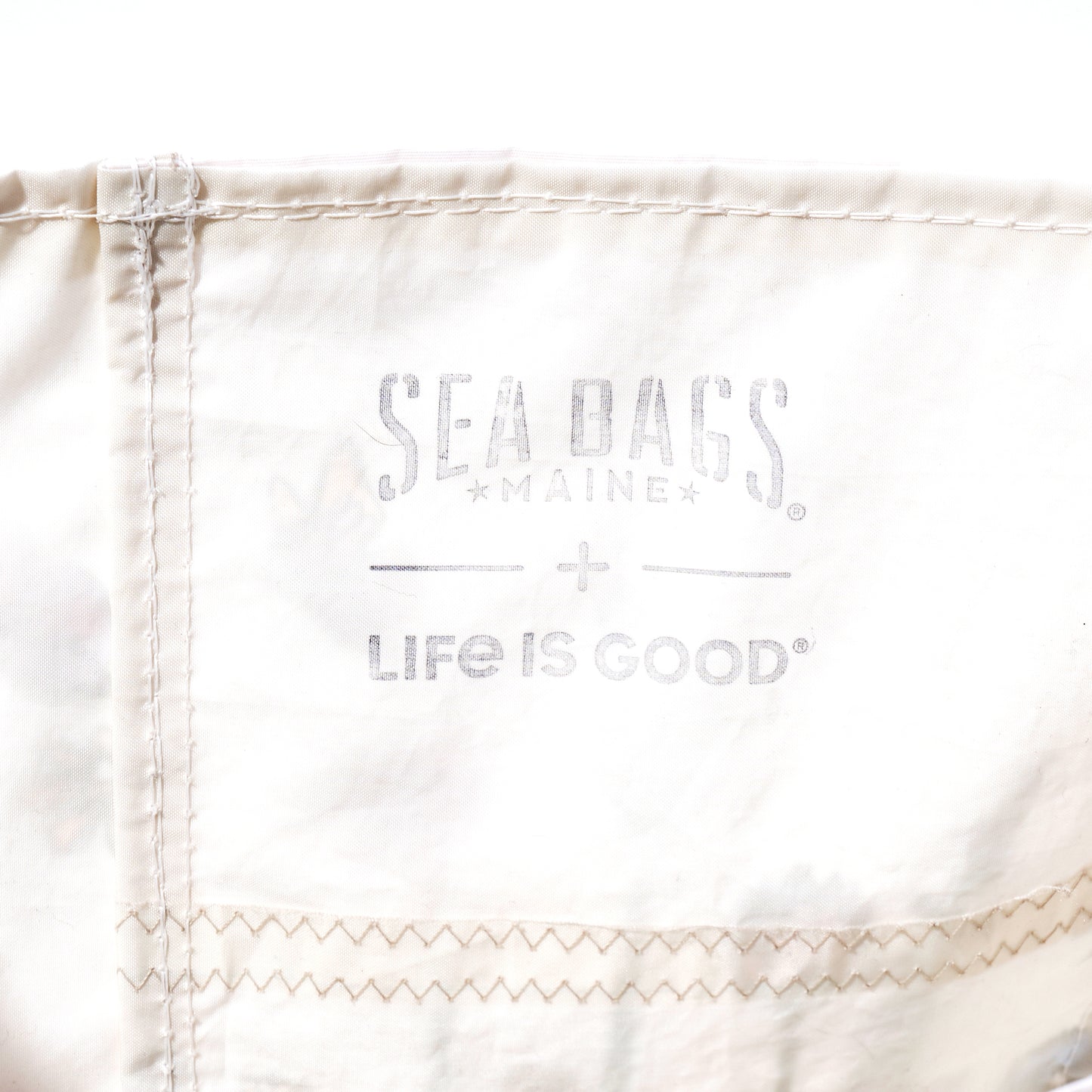 Handcrafted in Maine from recycled sail bag materials, the Life is Good Wildflowers Medium Tote by Sea Bags features white fabric with visible stitching and is printed with faded gray SEA BAGS MAINE and LIFE IS GOOD logos. by Sea Bags