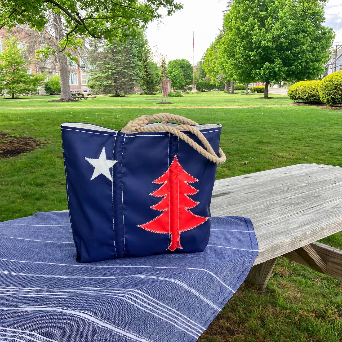 The Sea Bags 250th Patriotic Pine Medium Tote—a navy blue recycled sailcloth bag featuring a white star and red pine tree—rests on a blue-striped cloth atop a wooden picnic table in a grassy park. by Sea Bags