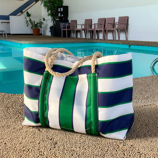 A Sea Bags Preppy Stripe Pier Tote, crafted from recycled sail cloth and featuring sturdy New England rope handles, sits on the ground beside an indoor swimming pool, with chairs and a potted plant in the background. by Sea Bags