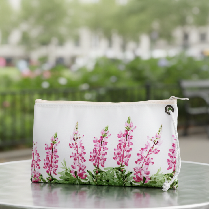 The Sea Bags Pink Lupine Large Wristlet, crafted from recycled sail cloth and featuring a pink lupine flower design, rests on a round metal outdoor table with greenery and a blurred park scene in the background. by Sea Bags