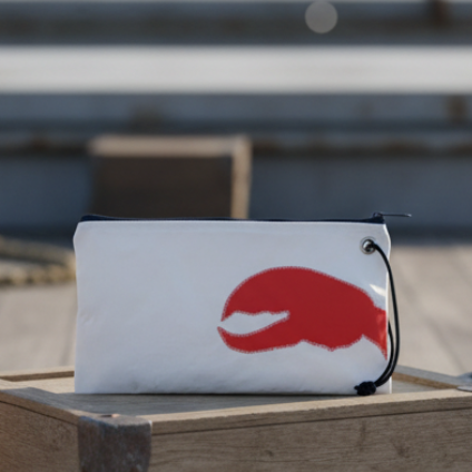 The Sea Bags Trapline Large Wristlet - Lobster Claw features a striking red lobster claw graphic on white, crafted from recycled sail material. This ocean-inspired accessory sits on a wooden surface with blurred steps in the background, offering nautical charm and eco-friendly style for your everyday essentials. by Sea Bags