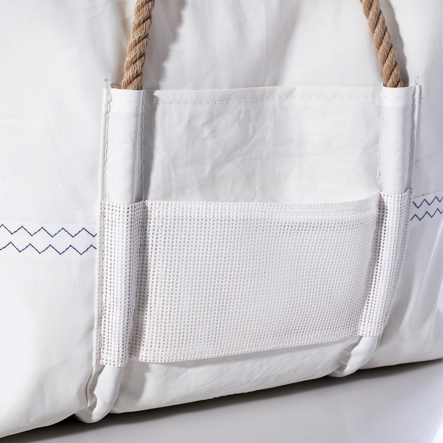 Close-up of the Sea Bags Trolley Sleeve Travel Tote, featuring white recycled sailcloth with visible blue zigzag stitching, tan rope handles, and a mesh front pocket with a trolley sleeve. by Sea Bags