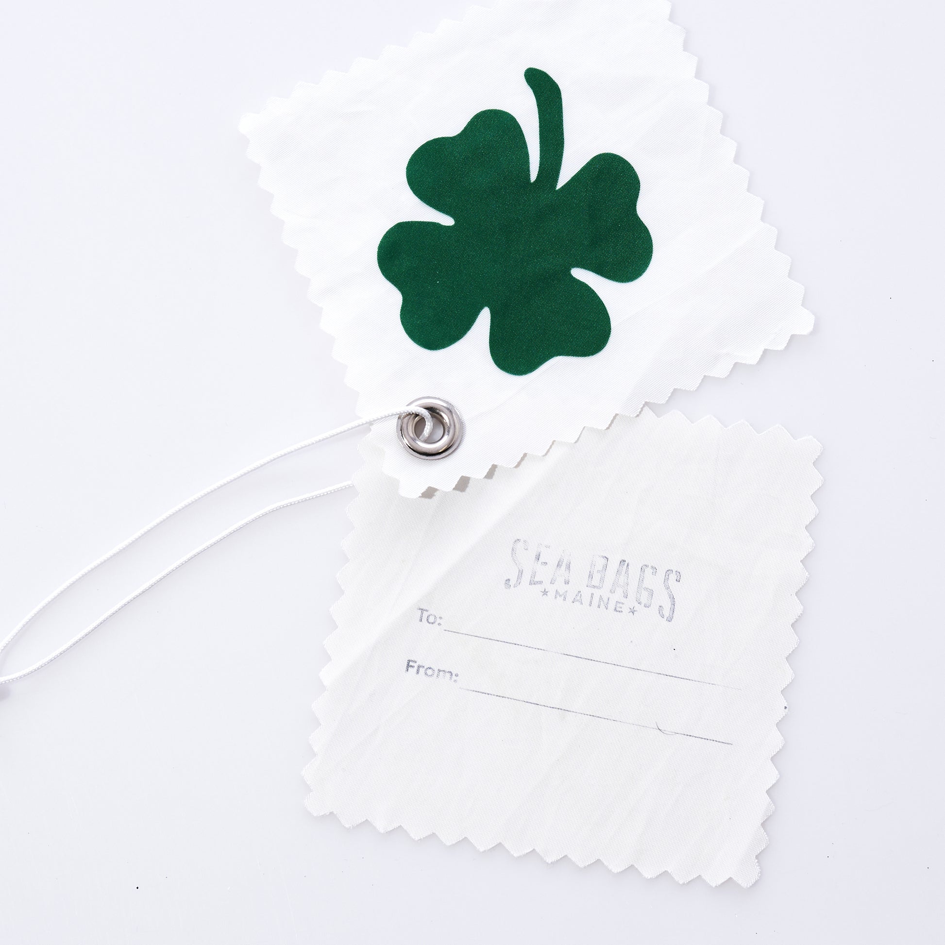 The Four Leaf Clover Sail Cloth Gift Tag by Sea Bags is crafted from white recycled sail cloth and features a green four-leaf clover design. Paired with a second tag labeled Sea Bags Maine and offering convenient To and From lines, both tags are joined by a sturdy white cord threaded through a metal grommet, making this accessory a sustainable way to personalize your gifts. by Sea Bags