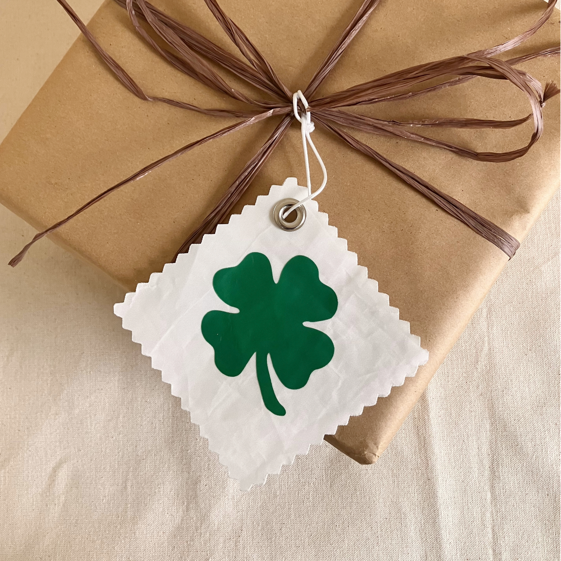 A gift wrapped in brown paper is adorned with brown raffia and the Sea Bags Four Leaf Clover Sail Cloth Gift Tag, a white tag featuring a green four-leaf clover—making it the perfect sustainable accessory for any occasion. by Sea Bags
