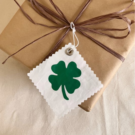 A gift wrapped in brown paper is adorned with brown raffia and the Sea Bags Four Leaf Clover Sail Cloth Gift Tag, a white tag featuring a green four-leaf clover—making it the perfect sustainable accessory for any occasion. by Sea Bags