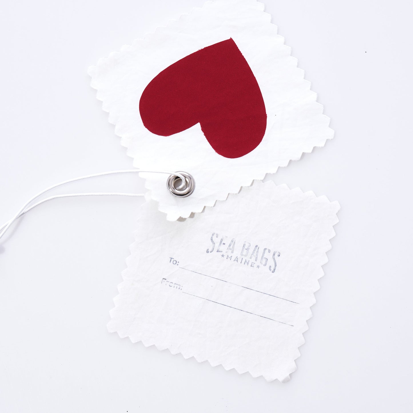 Introducing the Sea Bags Heart Sail Cloth Gift Tag—crafted from recycled sailcloth, this unique accessory features two white fabric tags: one adorned with a bold red heart and the other bearing the Sea Bags logo along with To: and From: fields. Connected by a sturdy metal grommet and string, this sustainable tag makes a thoughtful finishing touch for any gift or can serve as a distinctive charm on your bag. by Sea Bags