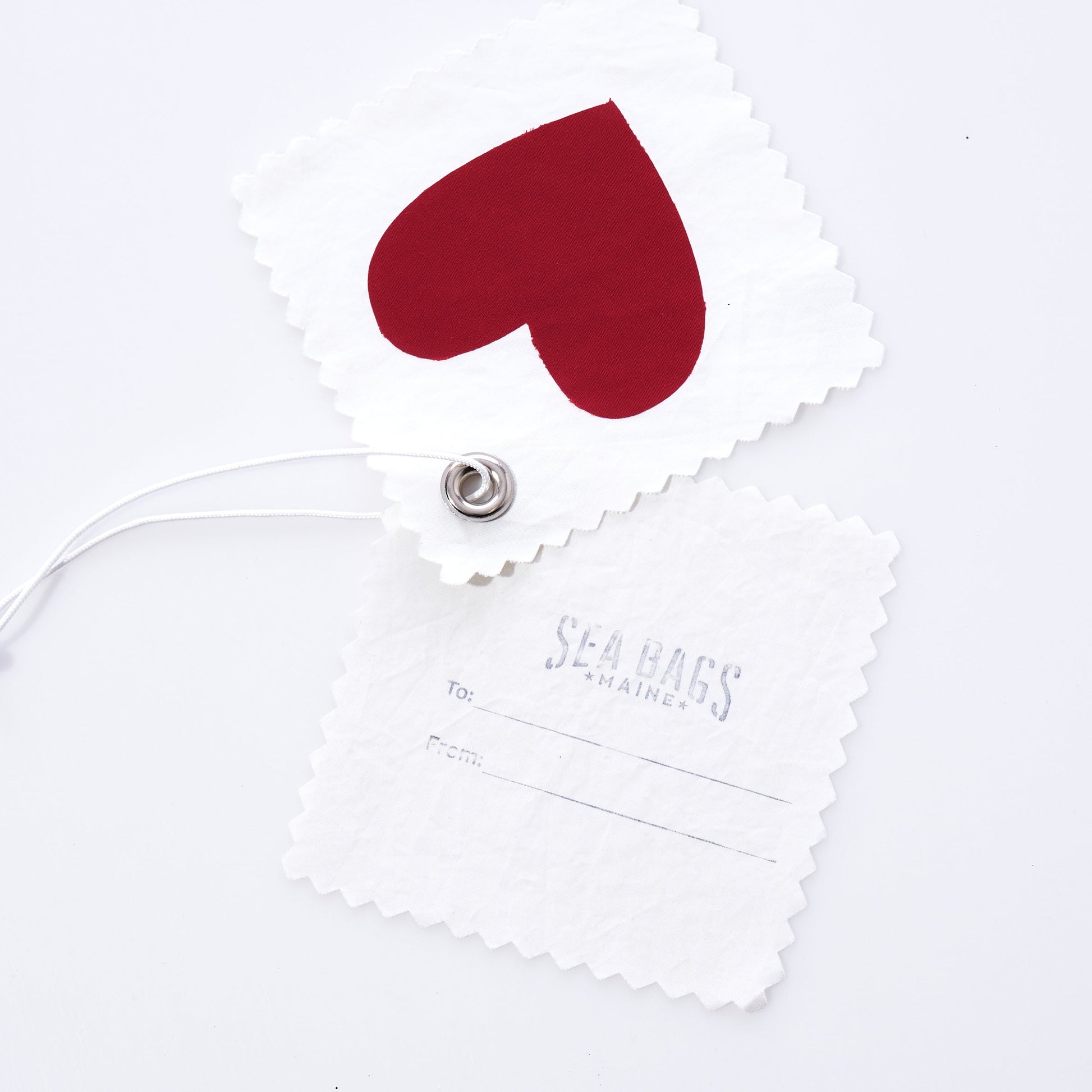 Introducing the Sea Bags Heart Sail Cloth Gift Tag—crafted from recycled sailcloth, this unique accessory features two white fabric tags: one adorned with a bold red heart and the other bearing the Sea Bags logo along with To: and From: fields. Connected by a sturdy metal grommet and string, this sustainable tag makes a thoughtful finishing touch for any gift or can serve as a distinctive charm on your bag. by Sea Bags