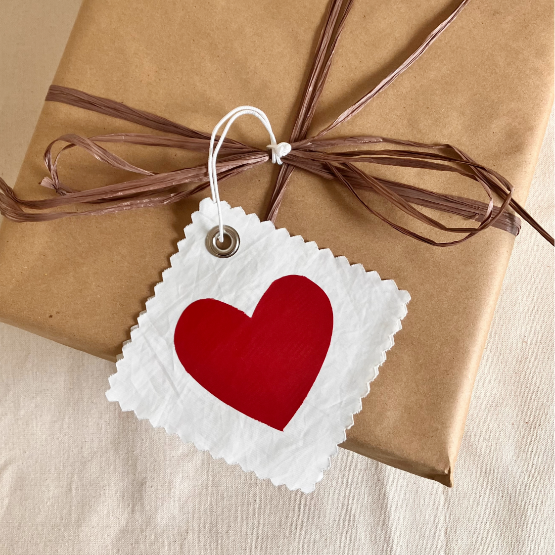 A brown gift box is tied with raffia ribbon and adorned with a Sea Bags Heart Sail Cloth Gift Tag, which features a large red heart on white recycled sail. by Sea Bags