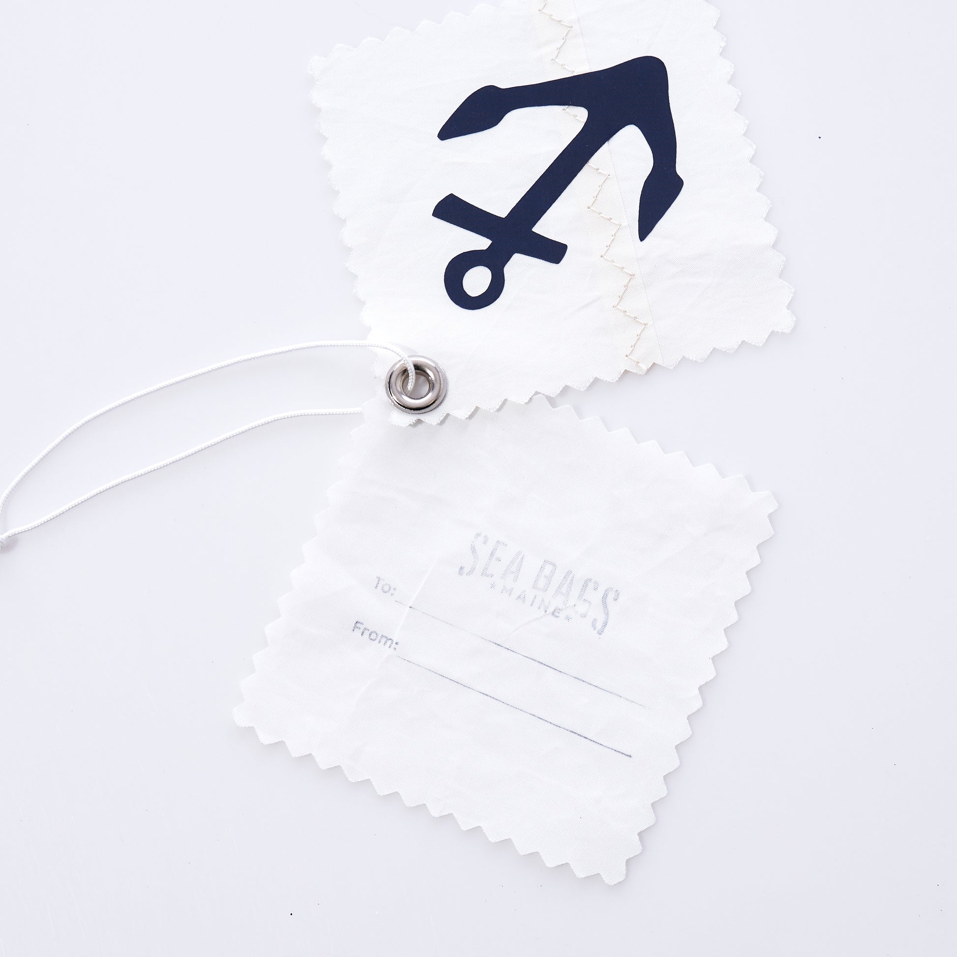 Two white fabric tags made from recycled sail cloth—one displaying a navy anchor graphic inspired by classic nautical motifs, and the other printed with subtle Sea Bags branding plus To/From fields—are joined by a white string and metal grommet against a white background. The Sea Bags Navy Anchor Sail Cloth Gift Tag provides an eco-friendly gift-wrapping solution with distinctive nautical style. by Sea Bags