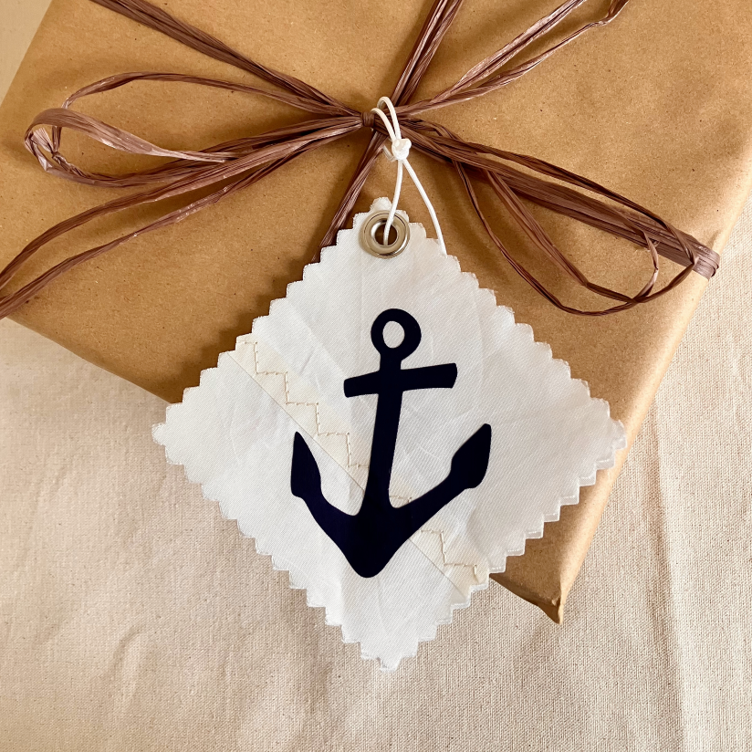A brown gift wrapped with twine features a Sea Bags Navy Anchor Sail Cloth Gift Tag—a white square recycled sail tag displaying a black anchor symbol. by Sea Bags