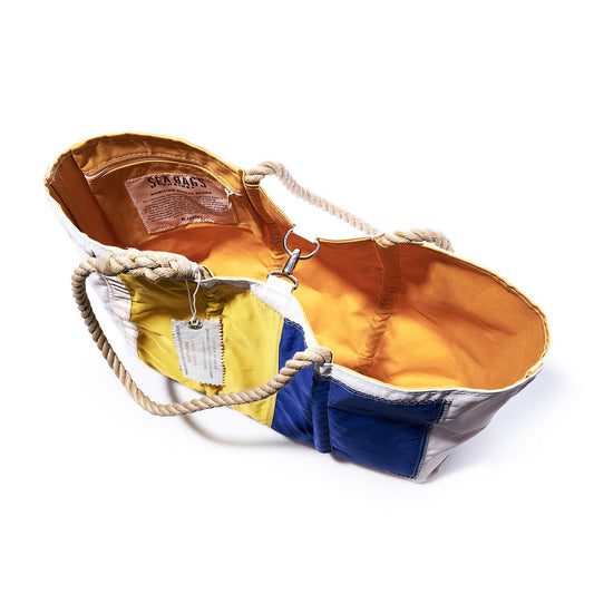 Introducing the Sea Bags Kilo Signal Flag Large Tote: an open maritime-inspired tote bag with sturdy rope handles, showcasing a patchwork exterior in blue, yellow, and white—a nod to vintage signal flags—with a vibrant orange lining. Expertly crafted from recycled sail cloth, this spacious tote features an interior label and secure metal clasp. by Sea Bags