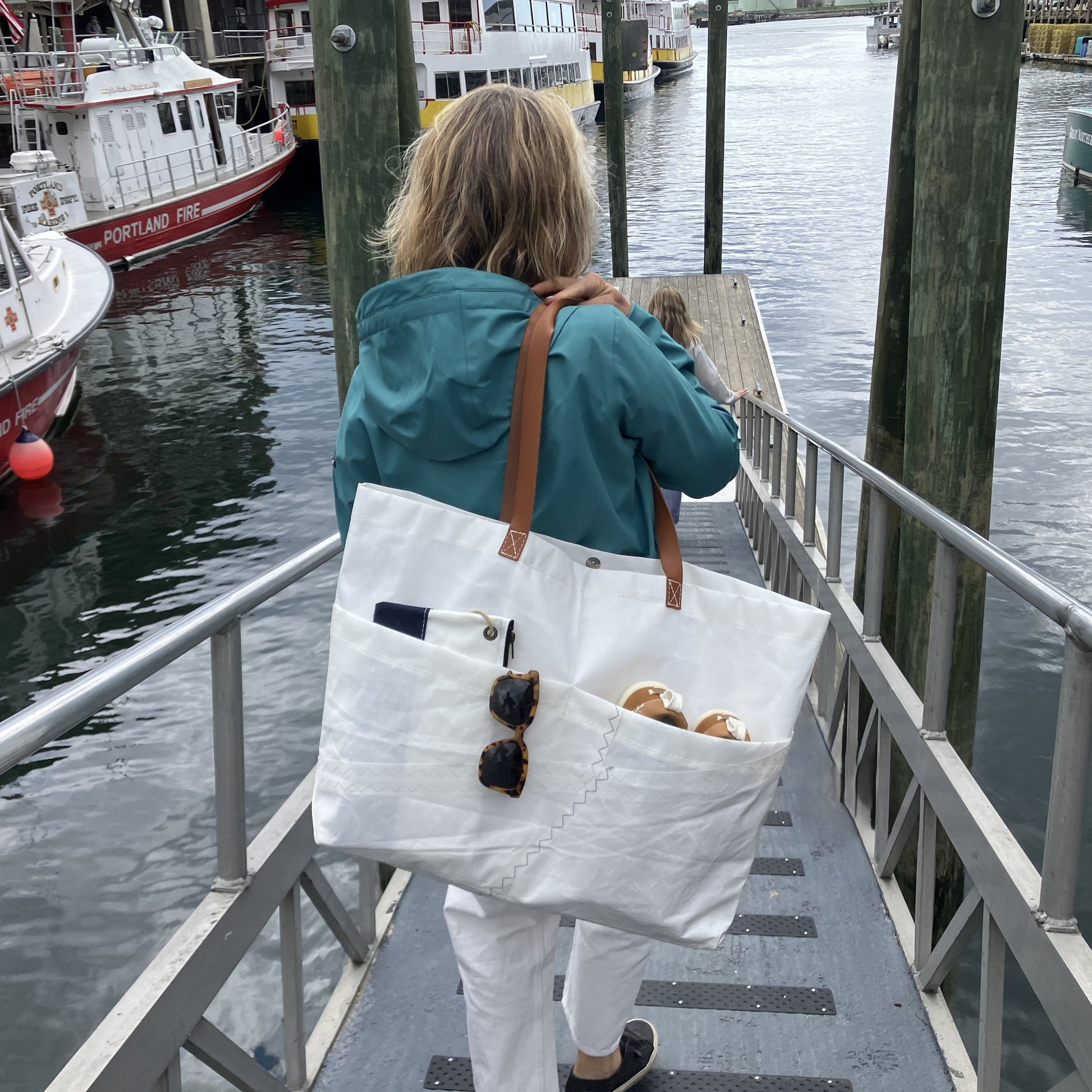 A person wearing a blue jacket carries the Sea Bags Large Market Tote—crafted from white recycled sail with leather handles—down a dock ramp toward boats; the spacious tote holds sunglasses, cans, and a phone. by Sea Bags