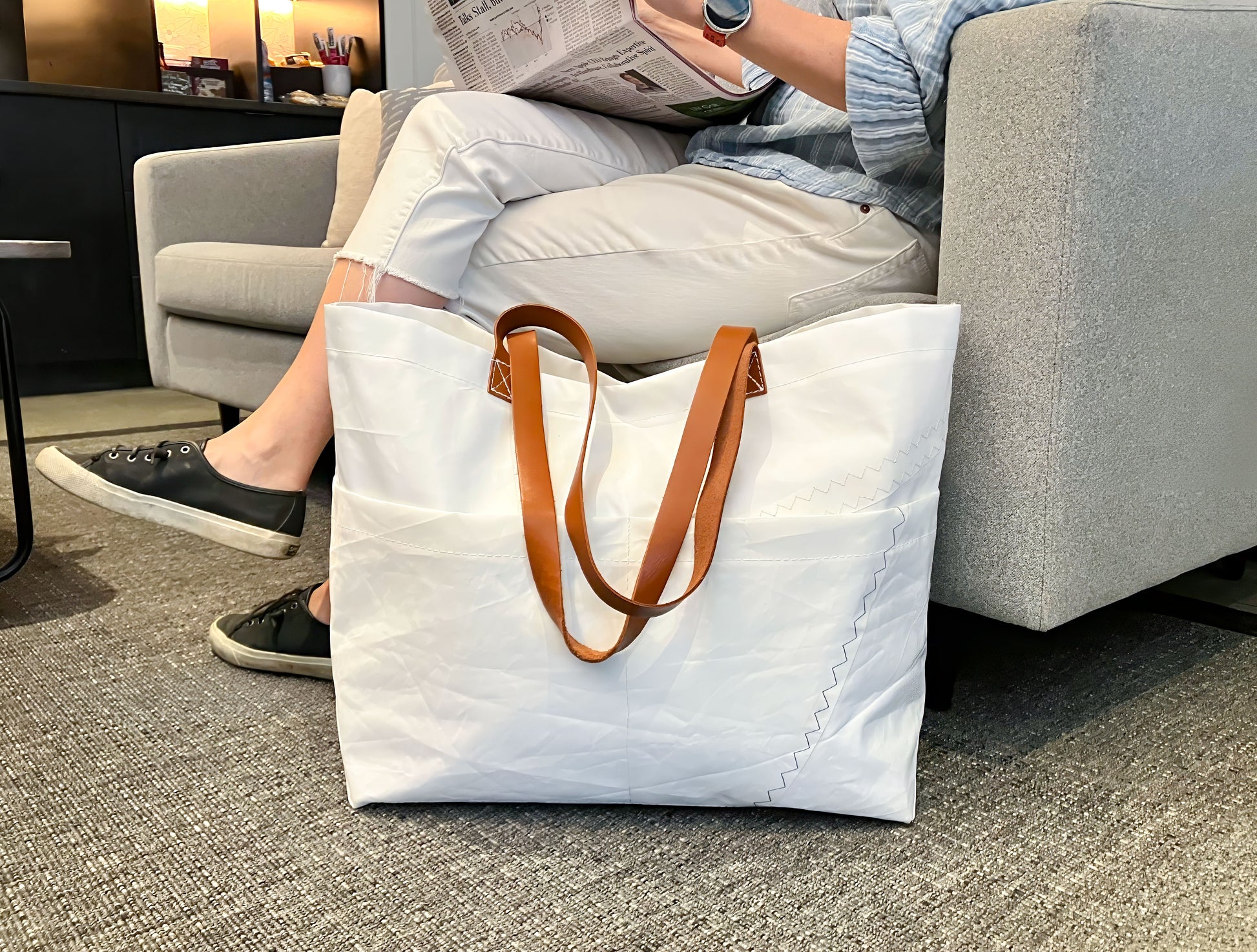 A person sits on a couch reading a newspaper, with the Sea Bags Large Market Tote—featuring brown leather handles—placed on the floor nearby. by Sea Bags