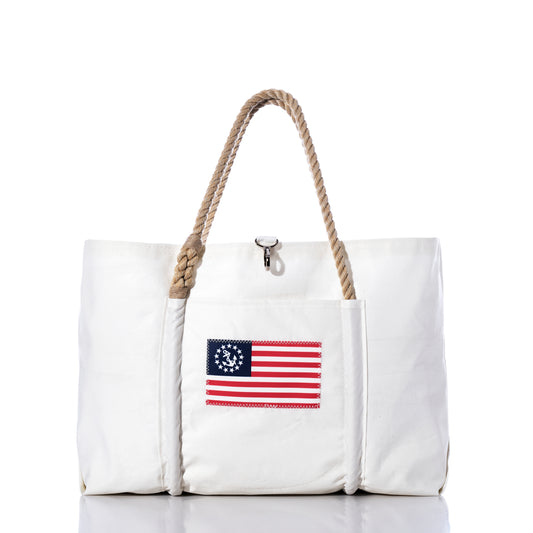 The U.S. Yacht Ensign Wharf Tote by Sea Bags is a white tote bag with rope handles, crafted from recycled sail cloth and featuring a vintage American flag patch with a circle of stars on the front pocket. Displayed against a plain white background, this piece blends sustainable craftsmanship with classic nautical style. by Sea Bags