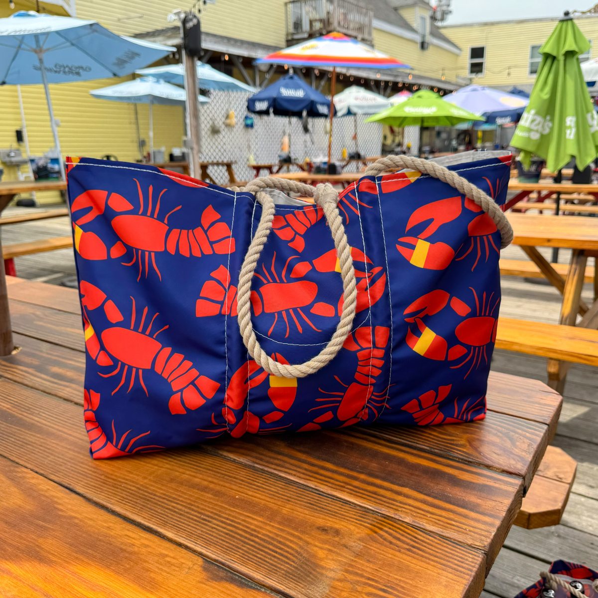 The Sea Bags Lobster Bake Large Tote, crafted from recycled sail cloth and adorned with red lobster prints and sturdy rope handles, sits on a wooden picnic table at an outdoor dining area with umbrellas. by Sea Bags