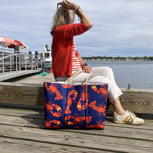 A woman in a red sweater and white pants sits on a dock by the water, holding her hair, with the Sea Bags Lobster Bake Large Tote—crafted from recycled sail cloth—beside her. by Sea Bags
