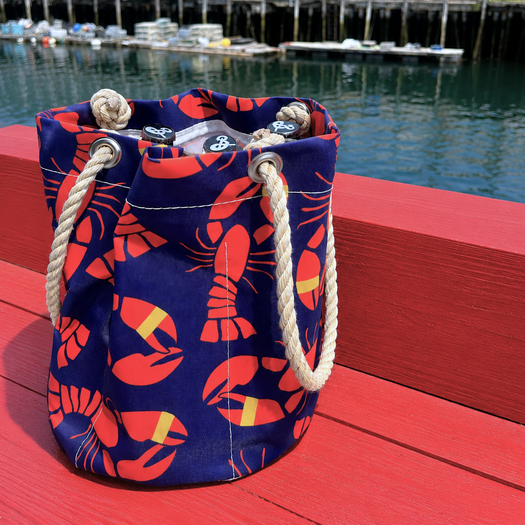 A Lobster Bake Beverage Bucket by Sea Bags, crafted from recycled sail cloth bag material and featuring blue drawstring accents with red lobster prints and rope handles, sits on a red bench by the water with docks and boats in the background—perfect for a day by the shore. by Sea Bags