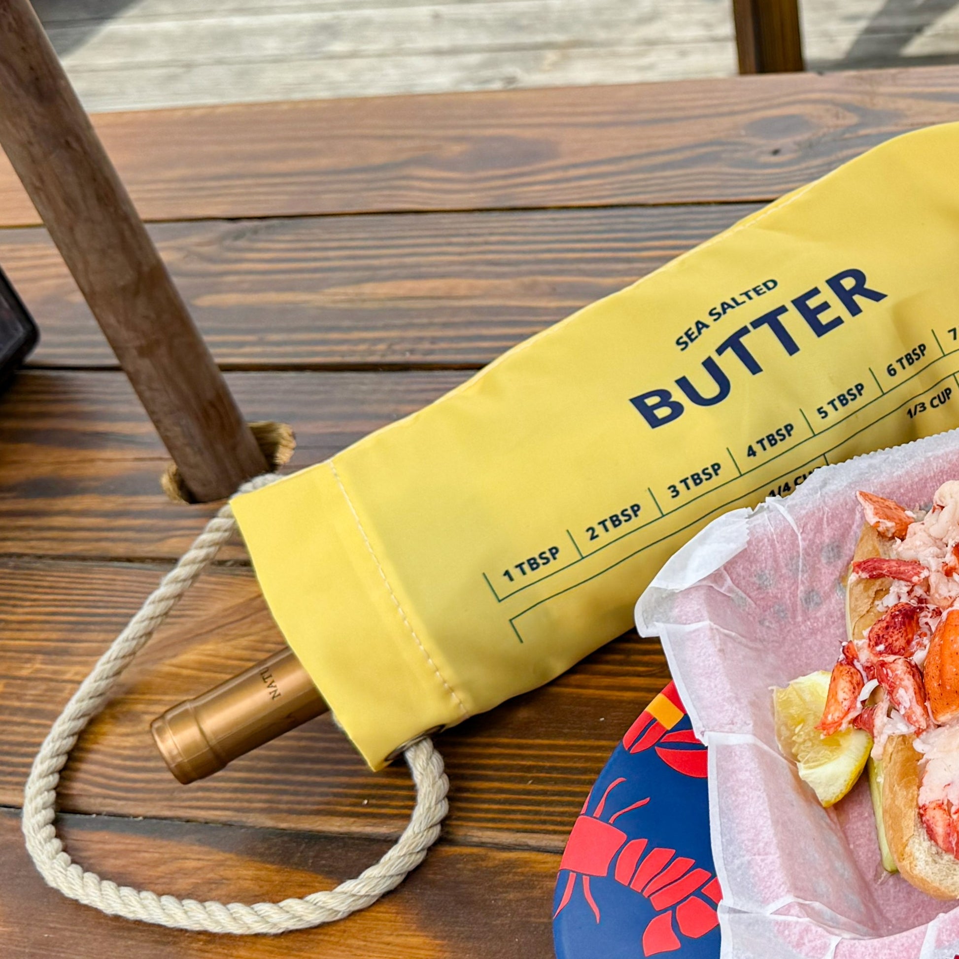 The Butter Wine Bag by Sea Bags, crafted from recycled sail cloth in a cheerful yellow butter motif, holds your favorite bottle and sits atop a wooden table beside a lobster roll with lemon wedges—an ideal accessory for your next lobster bake. by Sea Bags
