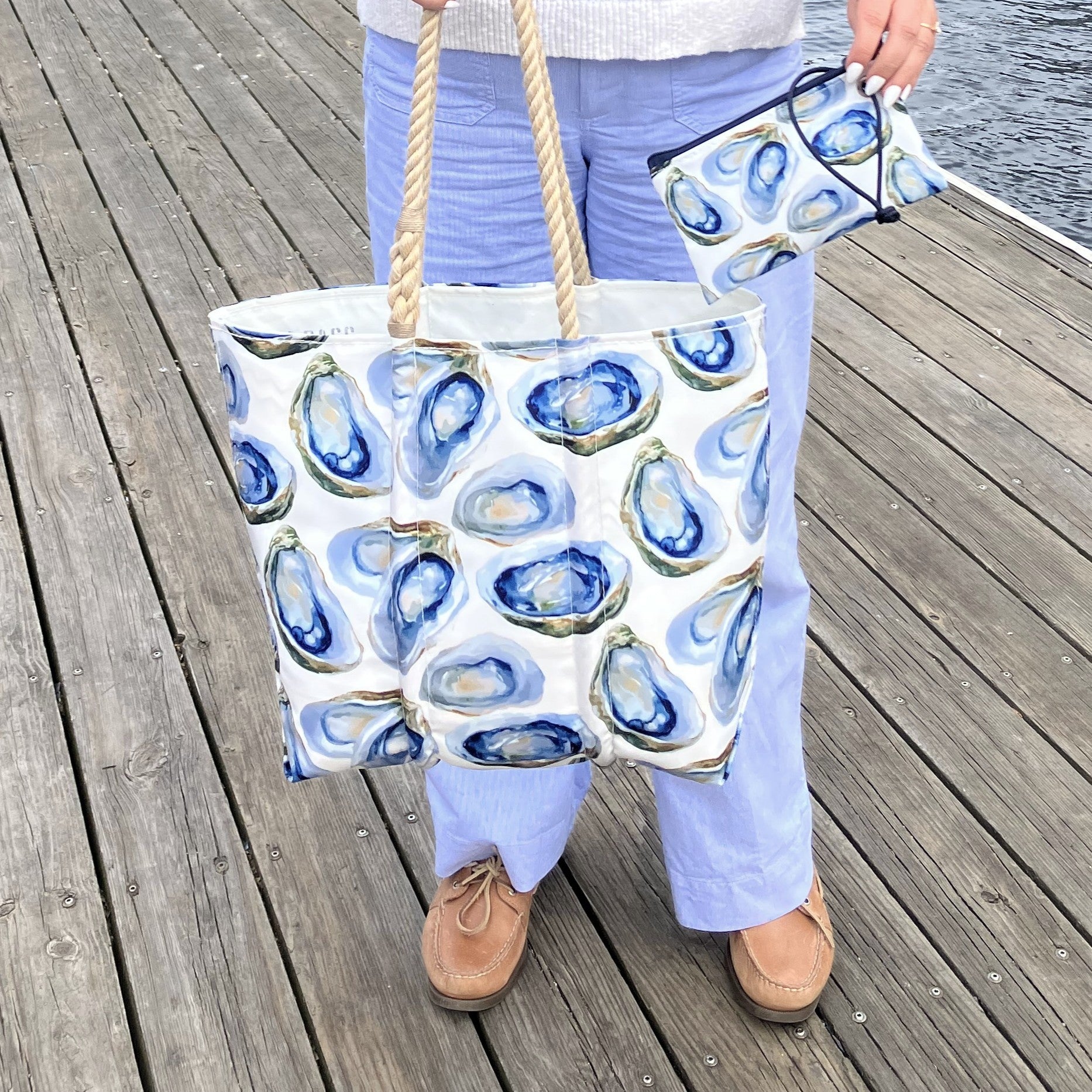 A person stands on a wooden dock holding the Sea Bags Watercolor Oyster Medium Tote and a matching pouch, both featuring blue oyster shell patterns. by Sea Bags