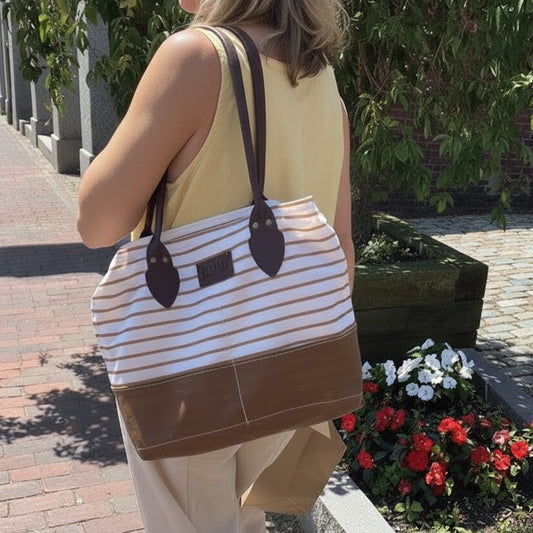 A person dressed in light-colored clothing strolls along a brick sidewalk near flowers, carrying the Sea Bags Tan Chebeague Stripe Handbag—a spacious, nautical-inspired bag made from recycled sail cloth and featuring brown handles—capturing the relaxed charm of Chebeague Island. by Sea Bags