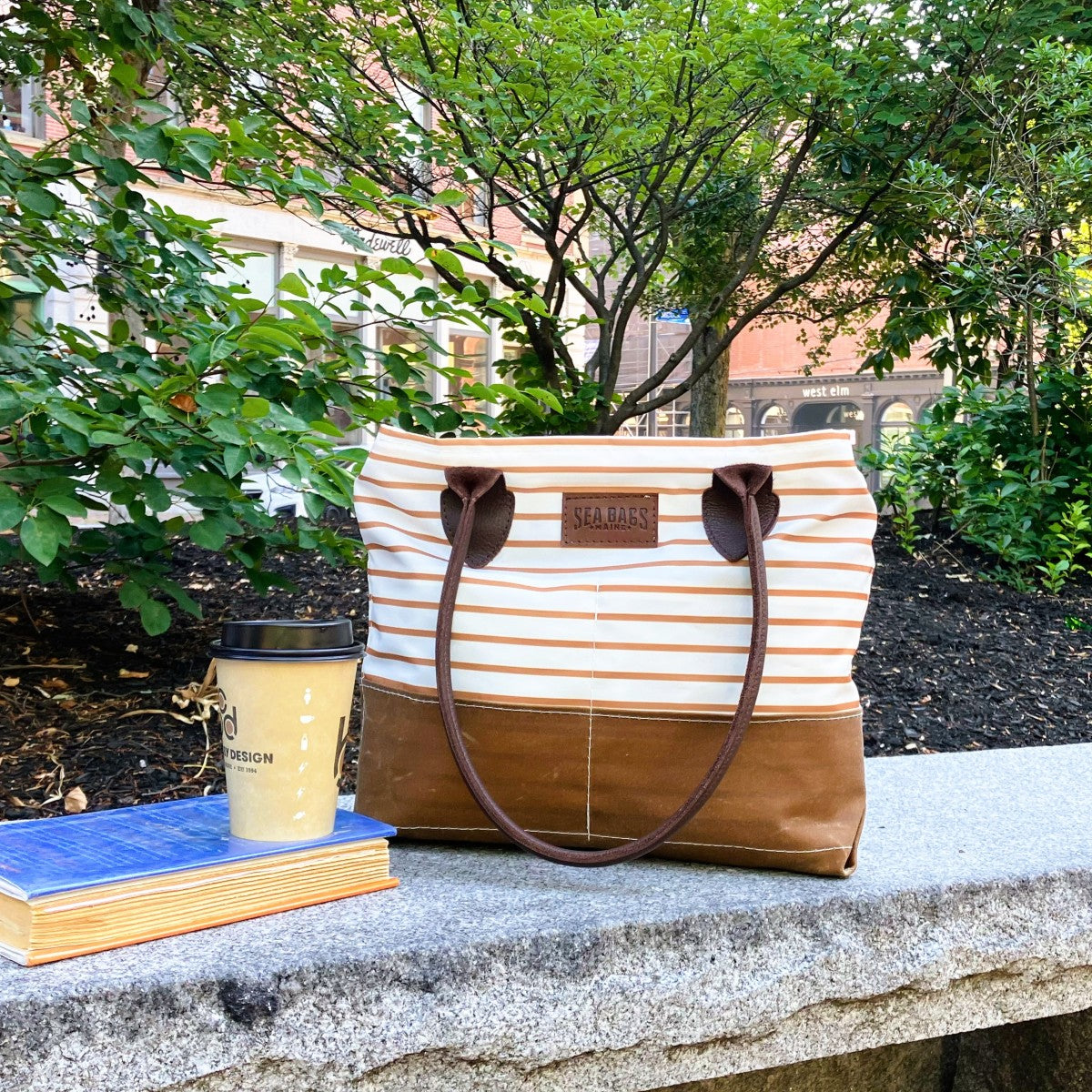 Here’s a rewritten version of the sentence, incorporating your product data: The Sea Bags Tan Chebeague Stripe Handbag rests on a stone bench alongside a stack of two books and a takeaway coffee cup in a landscaped outdoor setting with greenery and buildings in the background. by Sea Bags