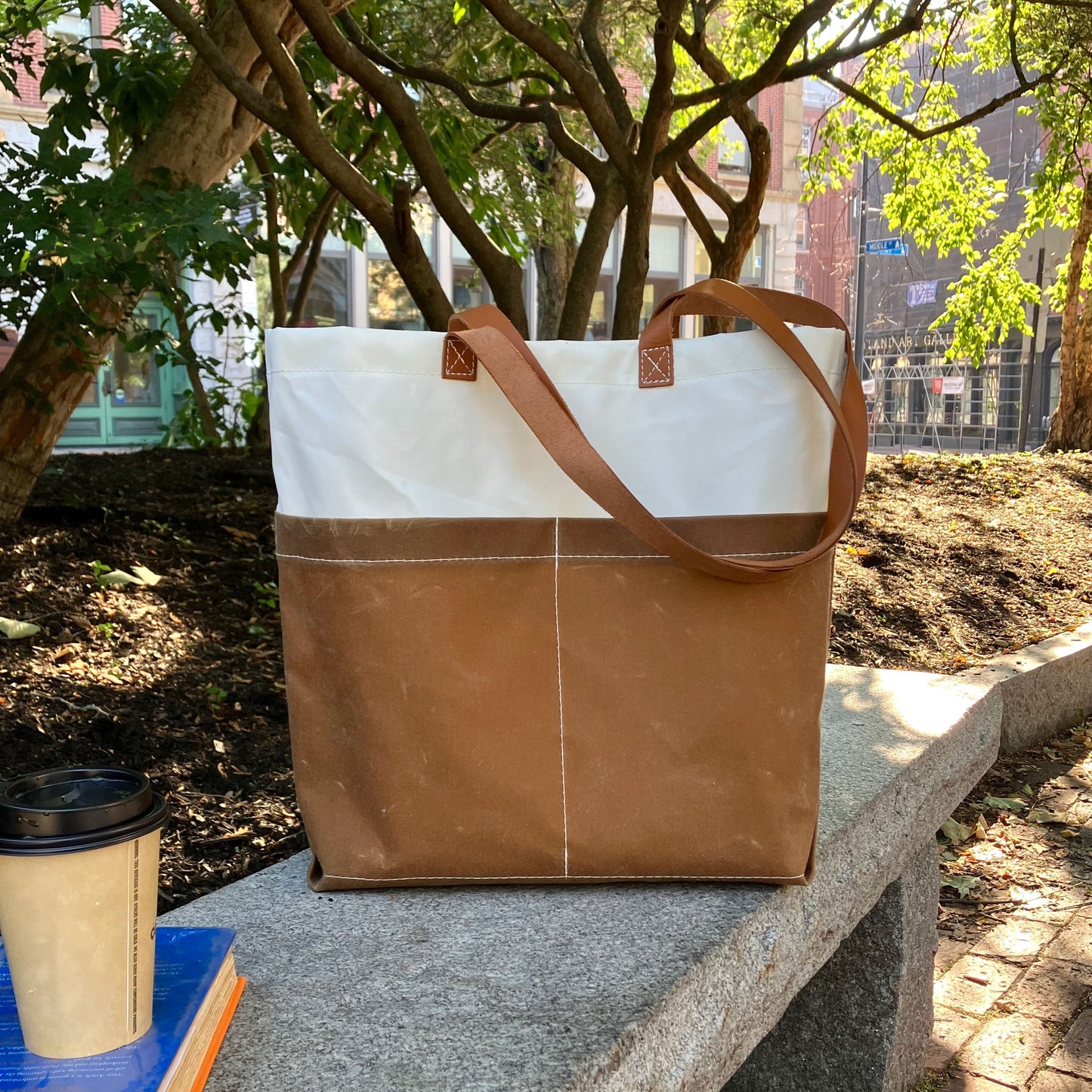 A Tan Chebeague Market Tote from Sea Bags, crafted from recycled sails, sits on a stone bench beside a coffee cup and a closed book, with trees and buildings in the background. by Sea Bags
