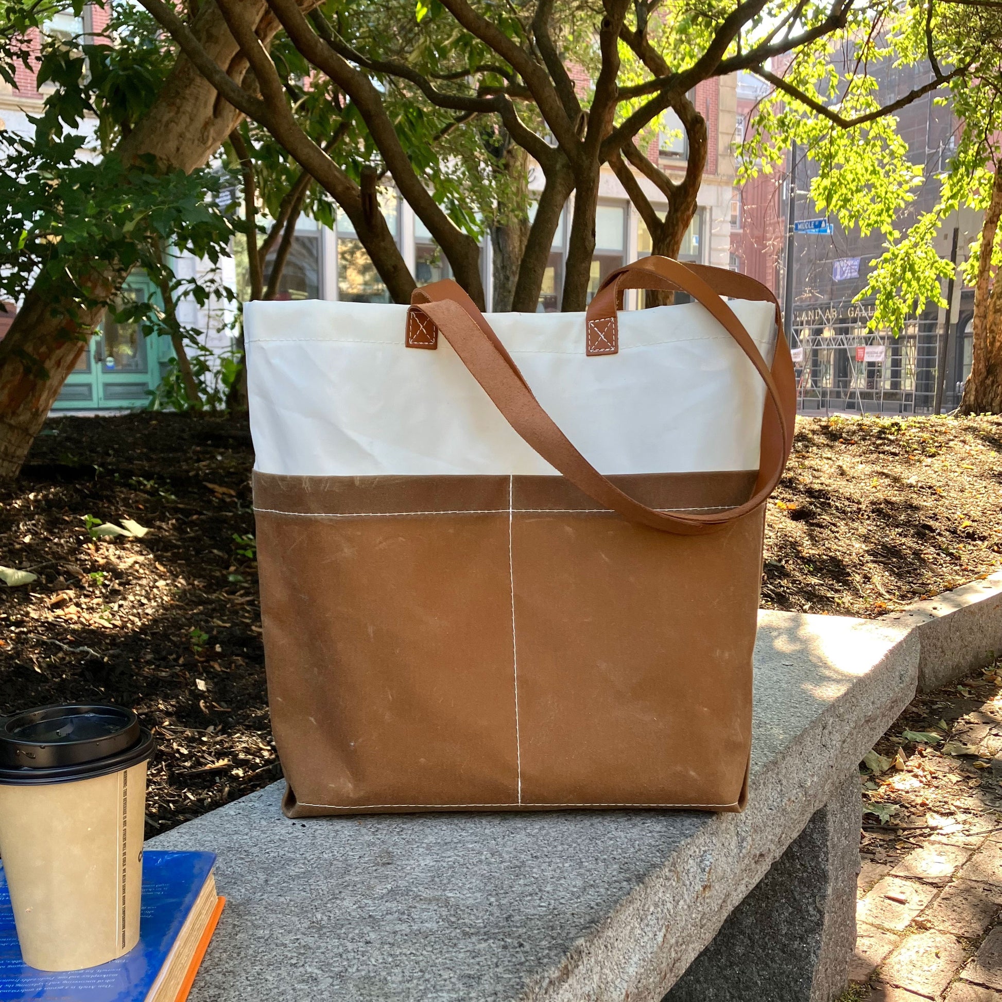 A Tan Chebeague Market Tote from Sea Bags, crafted from recycled sails, sits on a stone bench beside a coffee cup and a closed book, with trees and buildings in the background. by Sea Bags