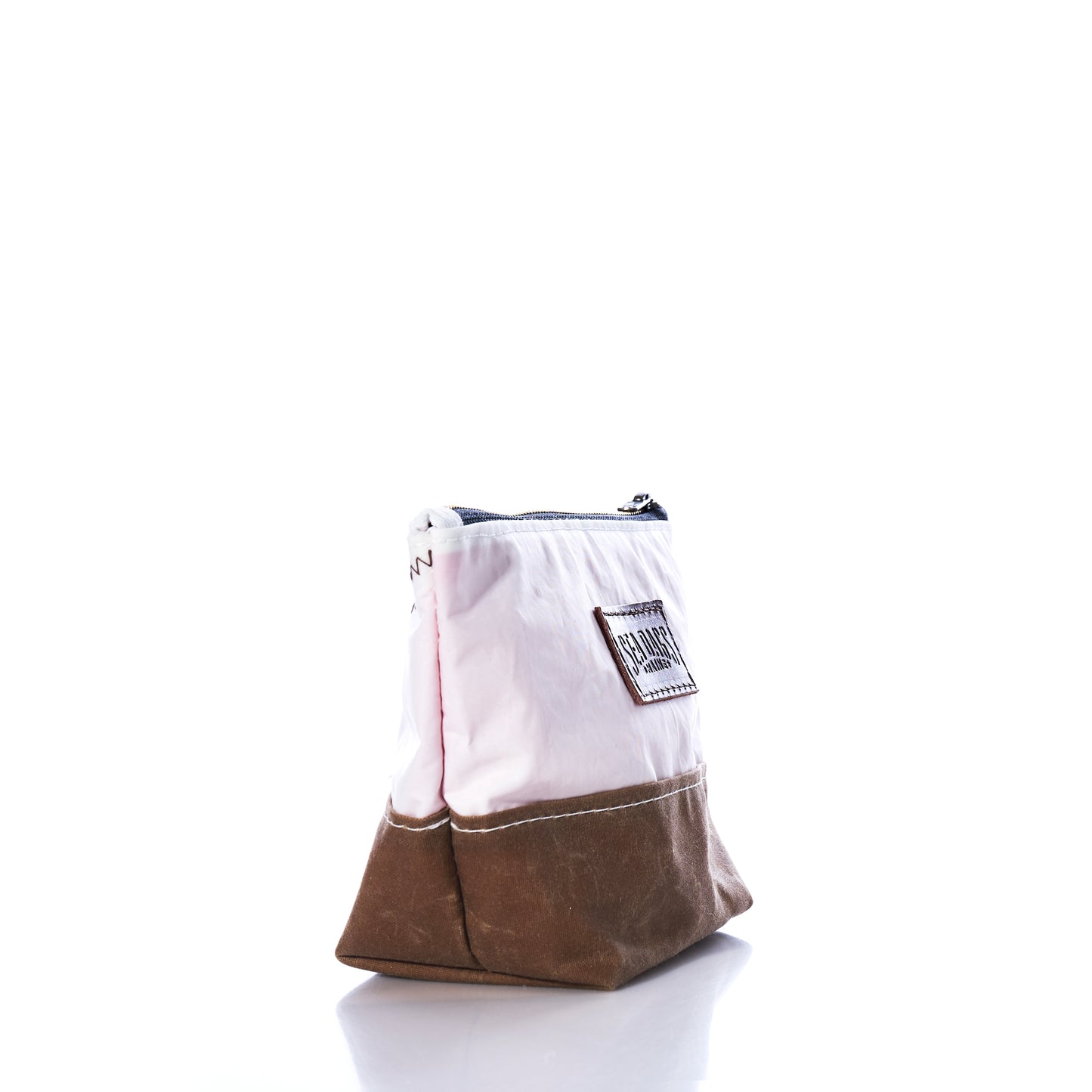 The Sea Bags Tan Chebeague Cosmetic Bag stands upright on a reflective white surface, showcasing its two-tone design crafted from recycled sailcloth with a white fabric upper half and tan base. Featuring a visible label and zipper closure, this sustainable accessory is perfect as a travel companion or cosmetic bag, blending eco-friendly materials with functional style. by Sea Bags